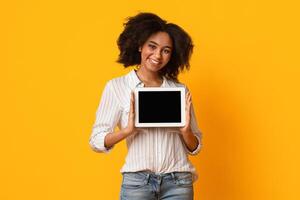 Cheerful Black Girl Showing Digital Tablet With Black Blank Screen, Holding Device In Her Hands And Sniling At Camera, Yellow Background, Mockup photo