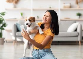 Happy young asian woman in casual sitting on floor in living room, taking selfie with her playful cute dog jack russel terrier puppy while playing together at home, using mobile phone, copy space photo