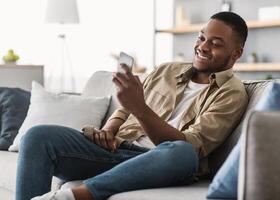 Side View Of African American Guy Using Smartphone Browsing Internet Sitting On Couch At Home. Black Man Texting On Cellphone Or Using New Application On Mobile Phone Indoors photo