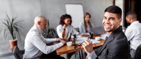 Young Businessman Smiling To Camera Sitting At Desk With Business Team During Corporate Meeting In Modern Office. Free Space photo