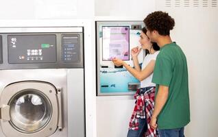 Two young adults stand close to a payment machine in a laundromat, reviewing options to start their laundry. They appear engaged as they navigate the machines interface. photo