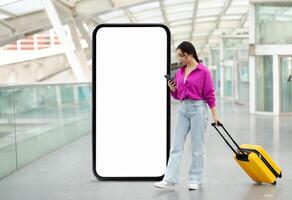 A traveler stands in a contemporary terminal, checking her smartphone while pulling a yellow suitcase. The spacious area features a large blank digital screen in the background. photo