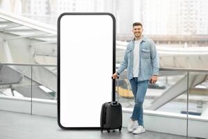 A young man stands beside a large blank display screen at a busy transportation hub, holding a suitcase. His casual attire contrasts with the sleek architecture around him. photo