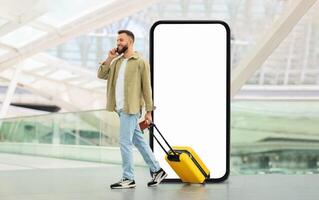 A traveler stands in a contemporary airport terminal, pulling a yellow suitcase while engaged in a phone conversation, passing by huge phone with blank screen photo