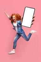 Happy young redhead woman jumping up, showing smartphone with empty screen, enjoying new application for mobile phone, full size photo. Pink studio background, creative image photo