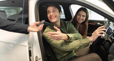 Happy millennial couple taking car key from auto salesman, sitting inside modern automobile at dealership, panorama. Cheery young family buying new vehicle at modern showroom photo