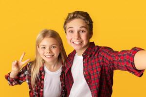 Brother and sister taking selfie, smiling and showing peace gesture while capturing photo together, yellow background, free space