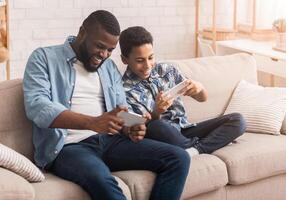 Happy Black Father And Son Playing Videogames On Smartphone Together, Competing With Each Other While Sitting On Couch In Living Room photo