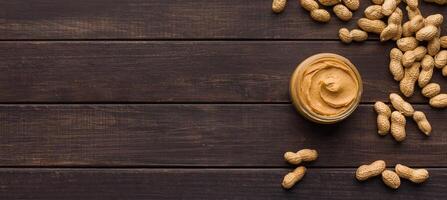 Bowl of peanut butter and scattered peanuts on dark wooden background, top view, copy space photo