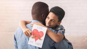 Little black boy embracing dad and giving him handmade postcard, congratulating with father's day while spending time together at home photo
