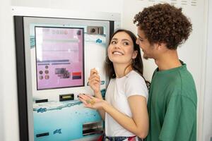 A smiling young woman holds snacks while her partner stands close, both enjoying a moment together at a vending machine in a well-lit area. photo