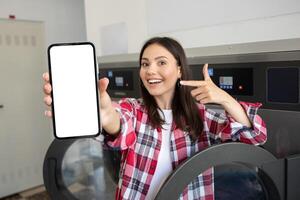 In a laundromat, a young woman happily points to her smartphone, displaying an empty screen, reflecting joy and engagement in her surroundings. photo