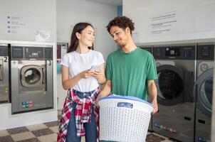 A young couple stands in a laundromat, smiling and engaged in conversation. The woman holds a smartphone, while the man carries a basket filled with clothes ready to be washed. photo