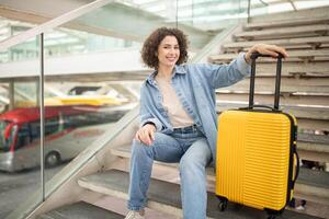 A cheerful young woman is seated on a staircase, smiling as she rests beside a bright yellow suitcase in a busy transportation hub, with buses in the background. photo