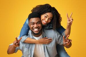Cheerful black lady piggybacking her boyfriend and showing peace sign while they posing together over yellow background in studio, free space photo