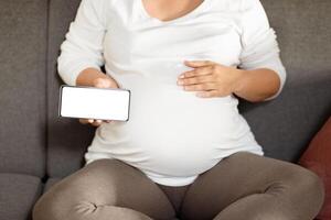 A pregnant woman relaxes on a couch, gently cradling her belly with one hand while holding her phone with the other. The atmosphere is calm, suggesting a cozy afternoon moment. photo