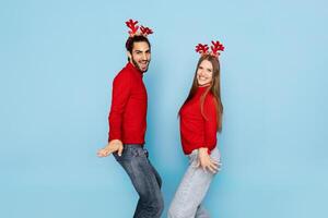 A cheerful couple in matching red sweaters and reindeer antlers poses playfully against a light blue background, exuding holiday spirit and joy during the festive season. photo