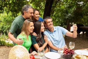 A cheerful group of five adults gather around a table outdoors, smiling and taking a selfie amidst greenery. They are enjoying a meal with grapes and drinks, creating joyful memories. photo