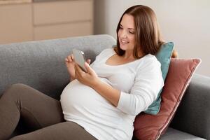 A pregnant woman sits comfortably on a couch, enjoying her time as she scrolls through her phone, surrounded by soft pillows in a cozy living space. photo