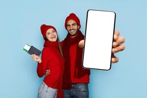 A joyful couple dressed in red winter clothing stands against a light blue background. The woman holds a passport, while the man displays a smartphone with a blank screen, smiling brightly. photo