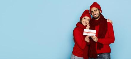 A cheerful couple is standing close together, both wearing cozy red sweaters and hats. They are smiling and holding a gift certificate in front of a light blue background, copy space photo