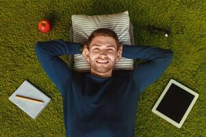Student Lifestyle. Relaxed Guy Lying On Green Grass Carpet Among Digital Tablet, Notebook And Eyeglasses. Top View photo