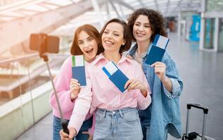 Three enthusiastic friends stand together at the airport, holding their tickets, smiling, and capturing the moment with a selfie stick. Their excitement for the upcoming journey is evident. photo