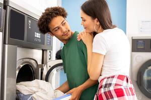 A young couple shares a moment at a laundromat, with one partner loading clothes into a washer while the other looks over affectionately, creating a warm and familiar atmosphere. photo