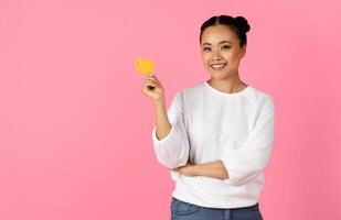 Asian young woman with a joyful expression displays a yellow card while standing against a vibrant pink backdrop. Her casual attire and playful pose suggest a fun, lively atmosphere. photo