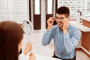 Choosing Eyewear Concept. Lady optometrist helping handsome man to pick correction eyeglasses, holding mirror photo