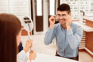 Eyewear Shopping Concept. Lady optometrist helping client to choose correction eyeglasses, holding mirror photo