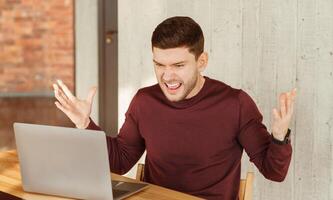 Angry Office Worker Shouting Sitting At Laptop Gesturing Having Problem With Computer At Workplace Indoor. Selective Focus photo