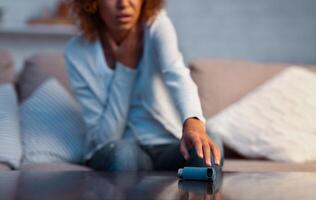 Afro girl suffering from asthma and taking inhaler from table, sitting on couch in living room photo