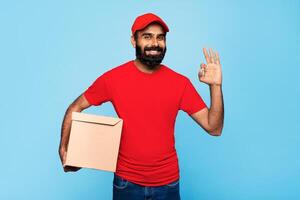 A bearded Indian man wearing a red cap and shirt is holding a brown box and smiling at the camera. He is making the okay sign with his right hand. photo