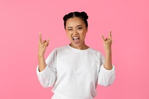 A young Asian woman is playfully making a rock gesture with her hands while winking and smiling. She stands out against a vibrant pink background, exuding confidence and joy. photo