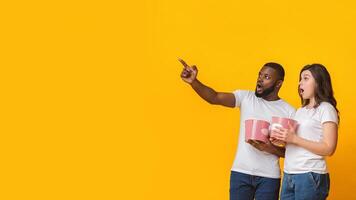 Scary movie. Shocked interracial couple holding popcorn buckets and pointing aside at copy space over yellow background, panorama photo