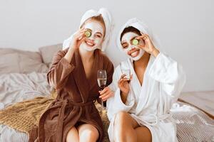 Cheerful multiracial girlfriends with face masks on wearing bathrobes drinking champagne, holding cucumber slices over eyes and smiling at camera, home interior photo