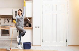 Stay At Home Routine. Happy woman in apron and yellow gloves waching floor, dancing with mop, free space photo