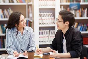 Two happy multiracial students talking at looking at each other sitting at desk in campus library, doing homework photo