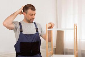 Puzzled Craftsman Scratching Head Assembling Shelf Set Without Manual Instruction Standing Indoors, Wearing Blue Coverall Uniform. Furniture Assembly Issues Concept photo