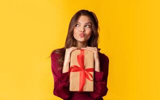 Portrait of funny lady with gift box pouting lips, grimacing and looking aside while posing over colorful background in studio, empty space photo