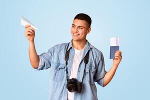 Playful guy traveler with camera holding passport, flight tickets and paper airplane, orange background photo
