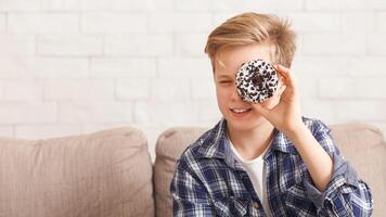 Boy Holding Doughnut Near Eye Posing Having Fun With Junk Food Sitting On Couch At Home. Panorama, Copy Space photo