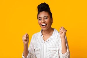 Joy Of Victory. Happy Black Woman Rejoicing Success, Shaking Fists Over Yellow Studio Background With Empty Space photo