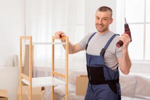 Furniture Assembler Man Posing With Electric Drill Assembling Cabinet Working Indoor, Smiling To Camera. Copy Space photo