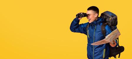 Surprised backpacker holding map, looking through binoculars at copy space, panorama, orange background photo