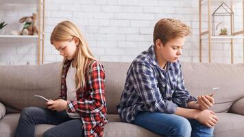 Brother And Sister Using Mobile Phones Sitting Back-To-Back Uninterested In Communication On Couch At Home. Modern Friendship photo
