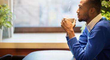 Good Start Of The Day. Pensive black guy holding white mug at cafe, drinking coffee at breakfast and thinking photo