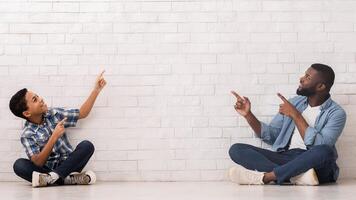 Free Space For Your Ad. Happy Black Father And Little Son Sitting On Floor Near White Brick Wall And Pointing At Empty Space photo