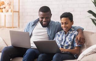 Common Hobby. Cheerful Black Father And Son Playing Games Online Together, Holding Laptops, Sitting On Sofa in Living Room photo